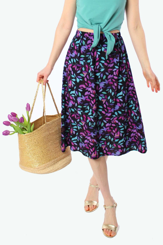 Midirock in Schwarz mit lila und türkisfarbenem Blütenmuster – luftiger, knielanger Sommerrock 2026 für Damen, kombiniert mit türkisfarbenem Top, goldenen Sandalen und Strohtasche.