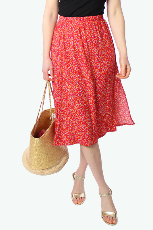 Detailaufnahme eines roten geblümten Midirocks aus Viskose mit feinem Blumenmuster, fließendem Stoff und leichtem Schlitz, kombiniert mit Strohtasche und goldenen Sandalen.