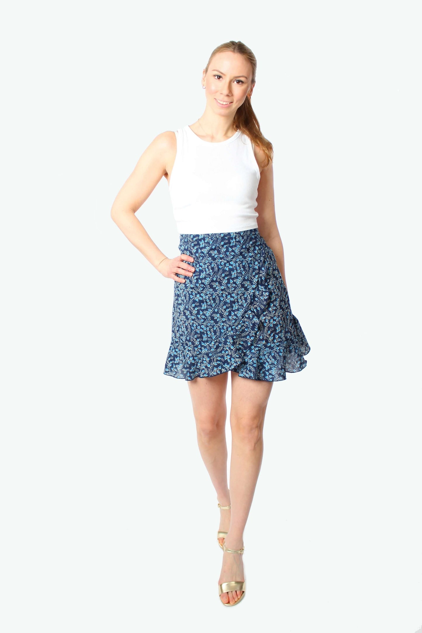 Blondes Model trägt einen kurzen dunkelblauen Wickelrock mit Blumenmuster und Volant, kombiniert mit weißem Top und Sandalen.