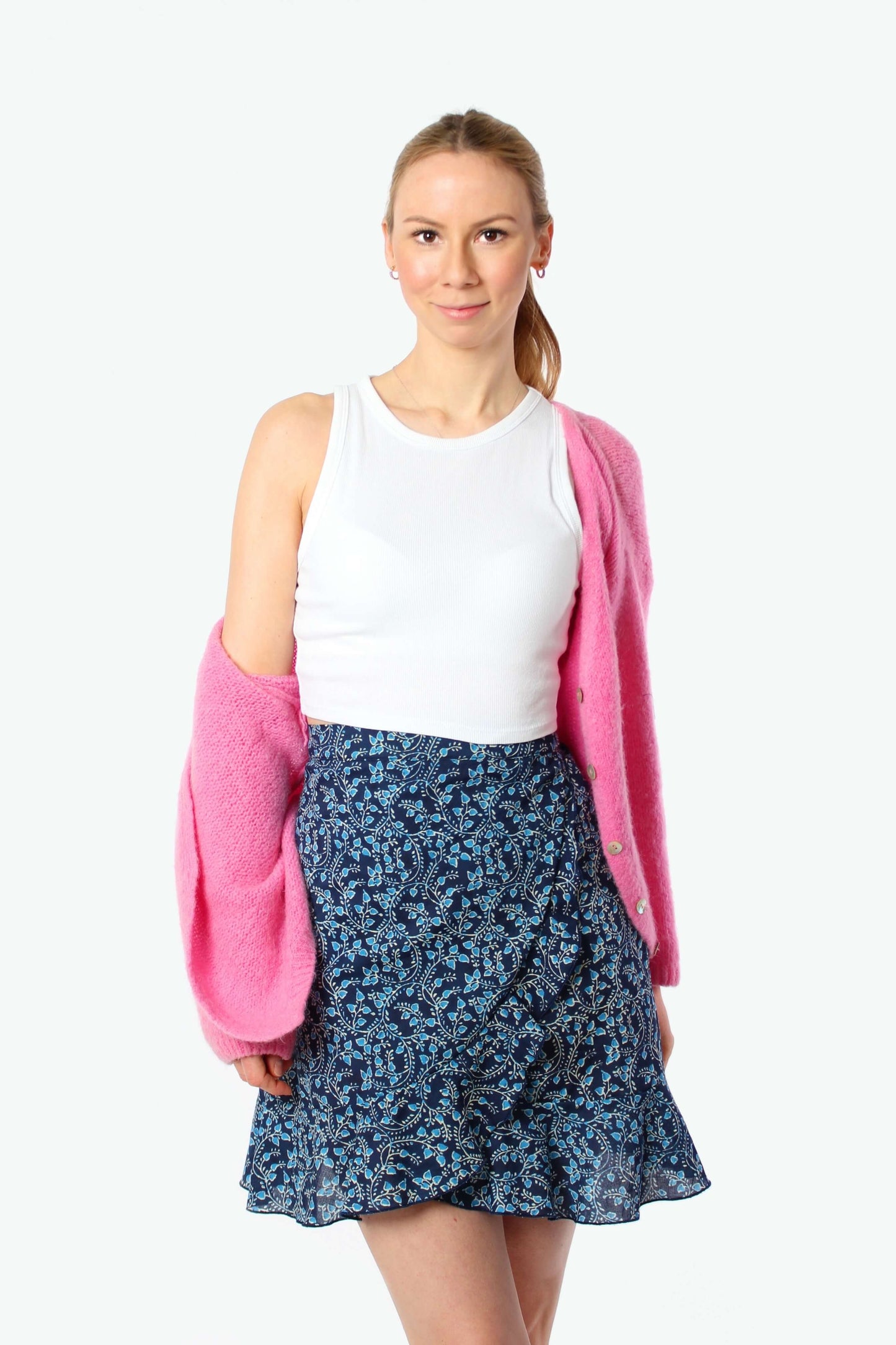 Blondes Model trägt einen dunkelblauen geblümten Wickelrock aus Baumwolle mit weißem Top und pinker Strickjacke.