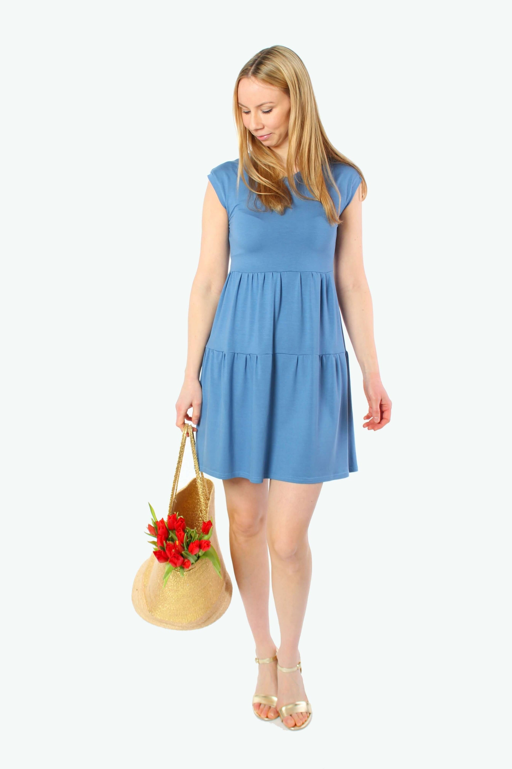 Model trägt hellblaues Sommerkleid in Minilänge mit kurzen Ärmeln und Stufenschnitt, locker fallendes Kleid, kombiniert mit Strohtasche und Sandalen.
