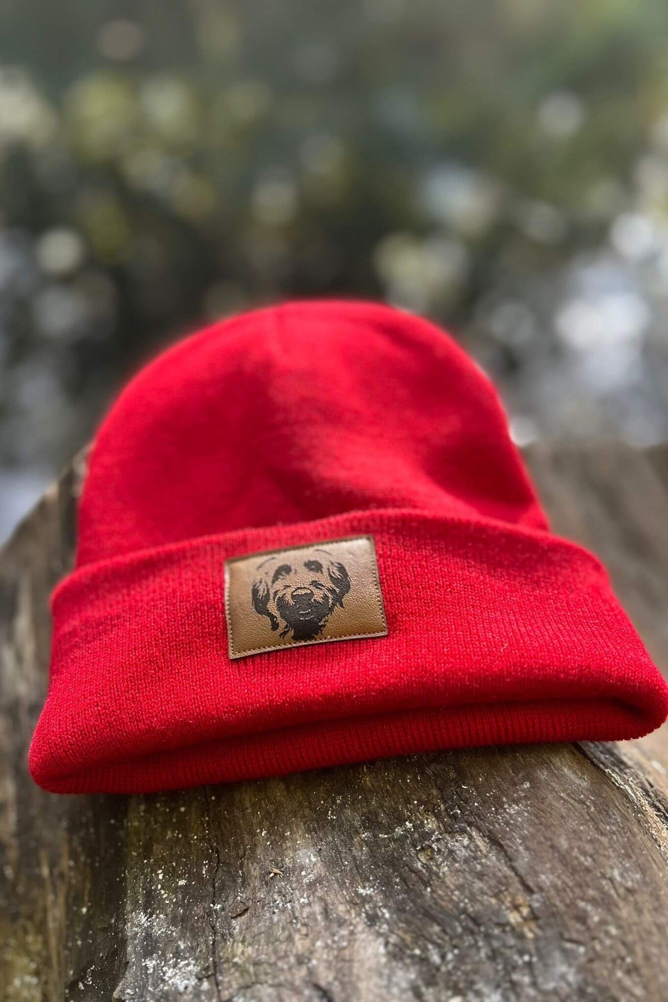 Rote Mütze von LA.LUKA Berlin mit braunem Kunstleder-Patch und Labradoodle-Motiv, platziert auf rustikalem Holz im natürlichen Licht – stylische Beanie für Hundefreunde.