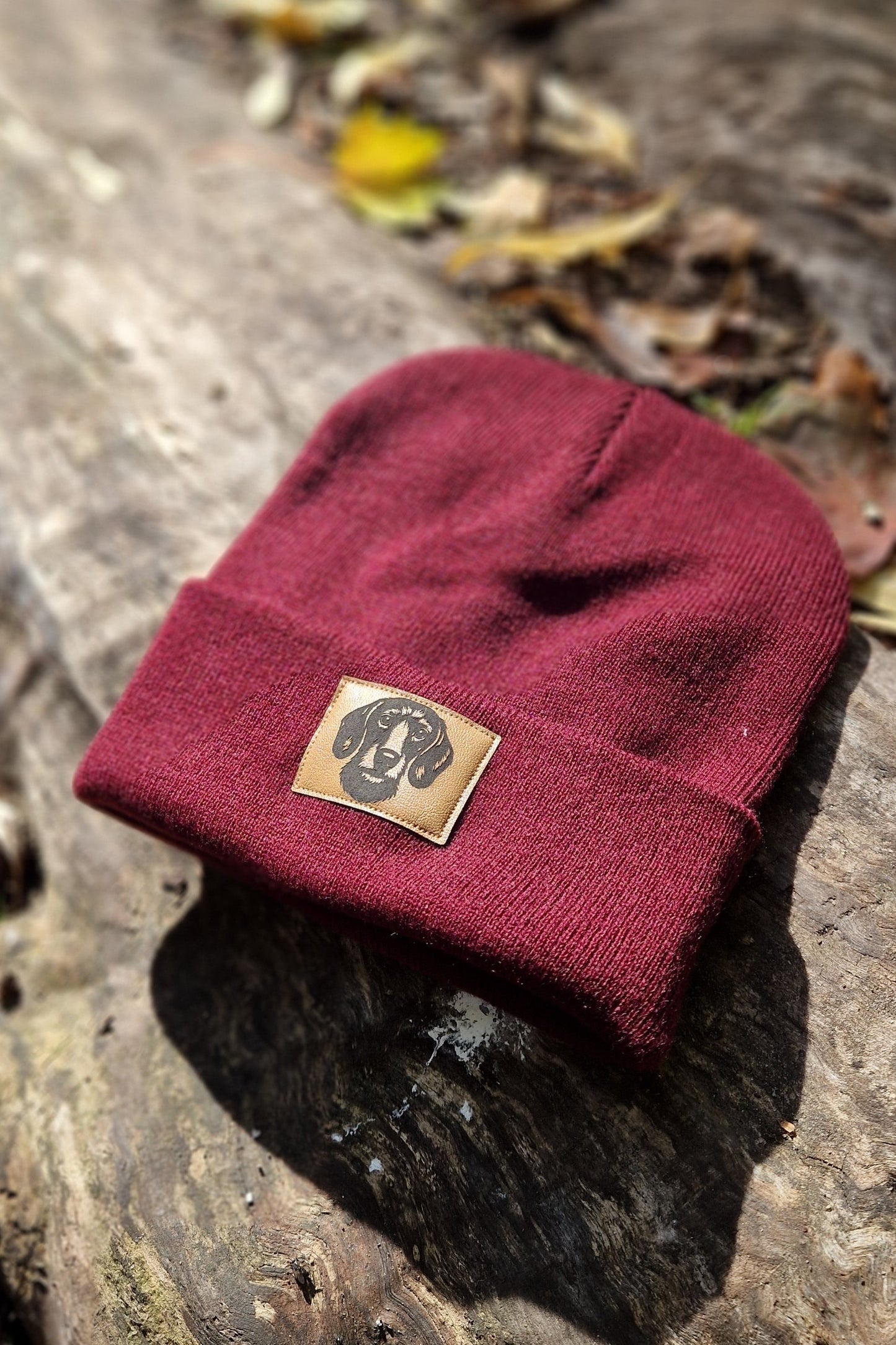 Bordeauxrote Mütze von LA.LUKA Berlin mit braunem Kunstleder-Patch und Dackelmotiv, stilvoll auf Holz mit Herbstlaub arrangiert – warme Beanie für Hundefans.