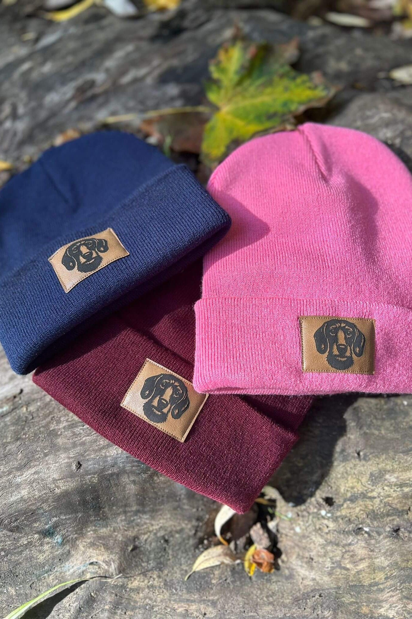 Drei Mützen in Bordeaux, Dunkelblau und Pink von LA.LUKA Berlin, jeweils mit Kunstleder-Patch und Dackelmotiv – farbenfrohe Beanies, perfekte Geschenkidee für Hundeliebhaber.