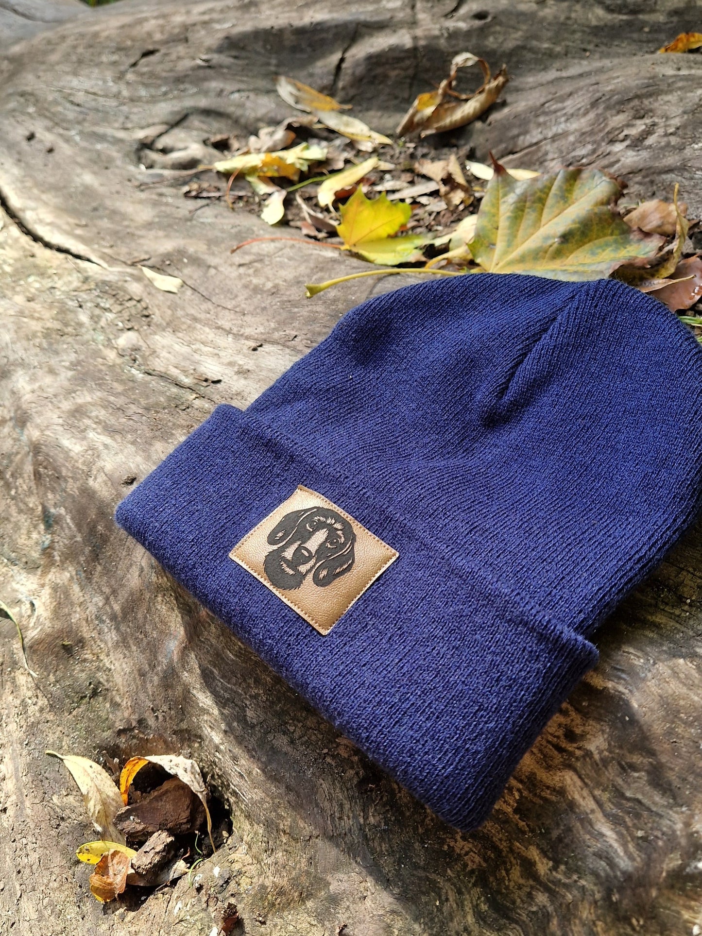 Dunkelblaue Mütze von LA.LUKA Berlin mit braunem Kunstleder-Patch und Rauhaardackel-Motiv, dekorativ auf Holz mit Herbstlaub fotografiert – stilvolle Beanie und ideales Geschenk für Hundeliebhaber.