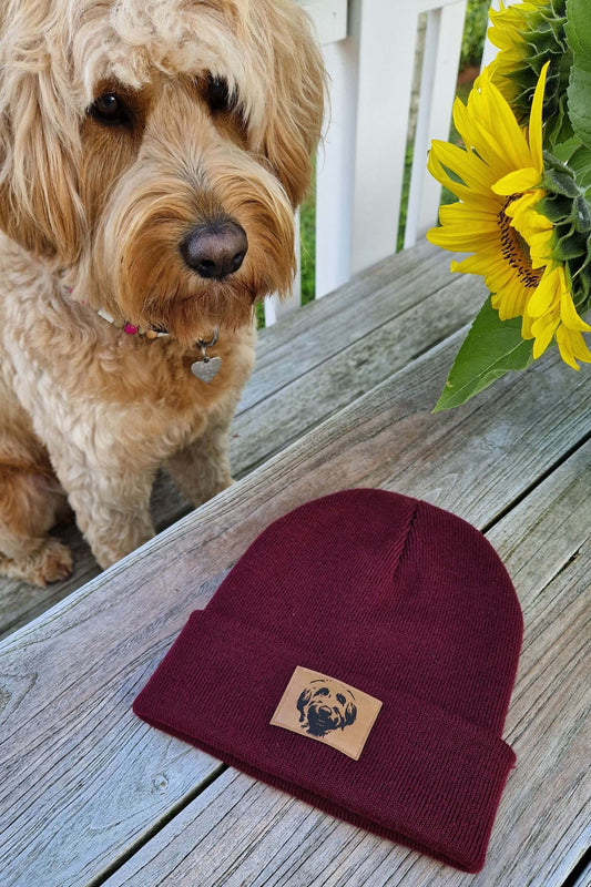 Mütze in bordeaux aus Feinstrick mit braunem Kunstleder-Aufnäher und Labradoodle-Motiv auf einem Holztisch. Im Hintergrund sitzt ein Labradoodle.