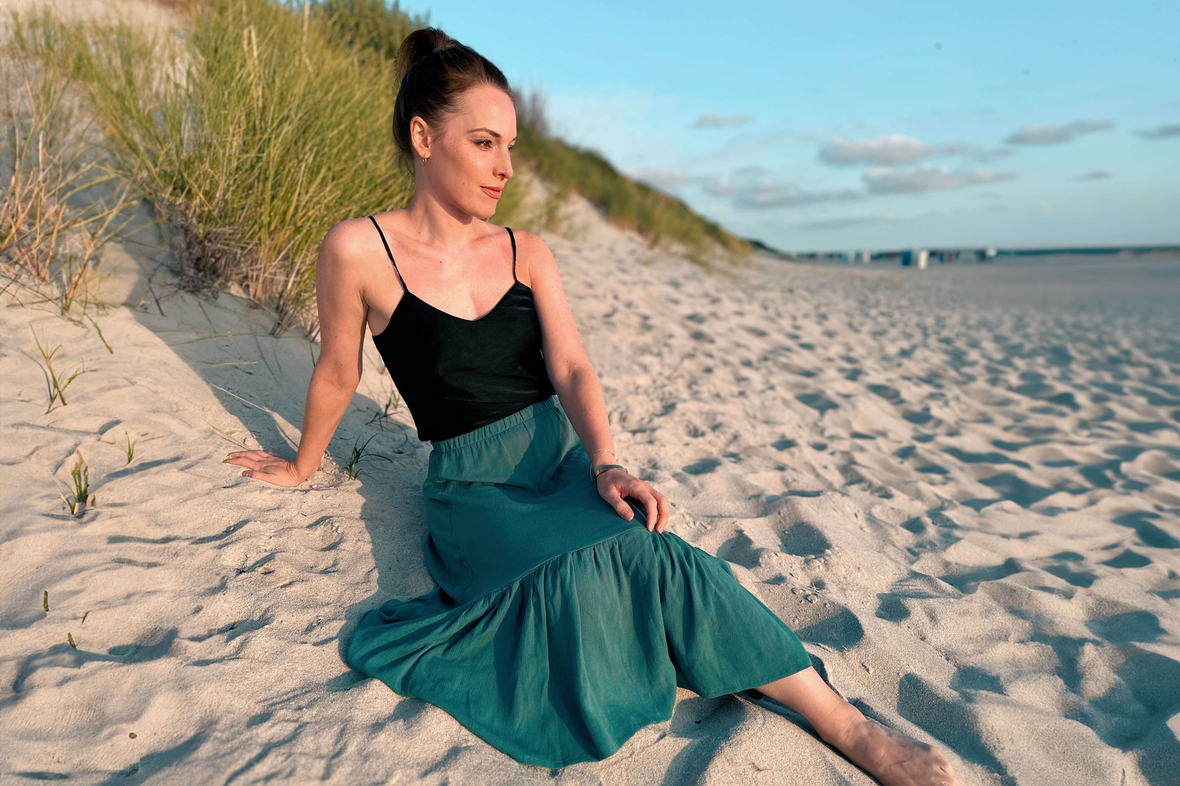 Frau, sitzt an einem Strand bei den Dünen. Sie blickt auf das Meer und trägt einen grünen Midirock von LA.LUKA Berlin mit einem schwarzen Trägertop.