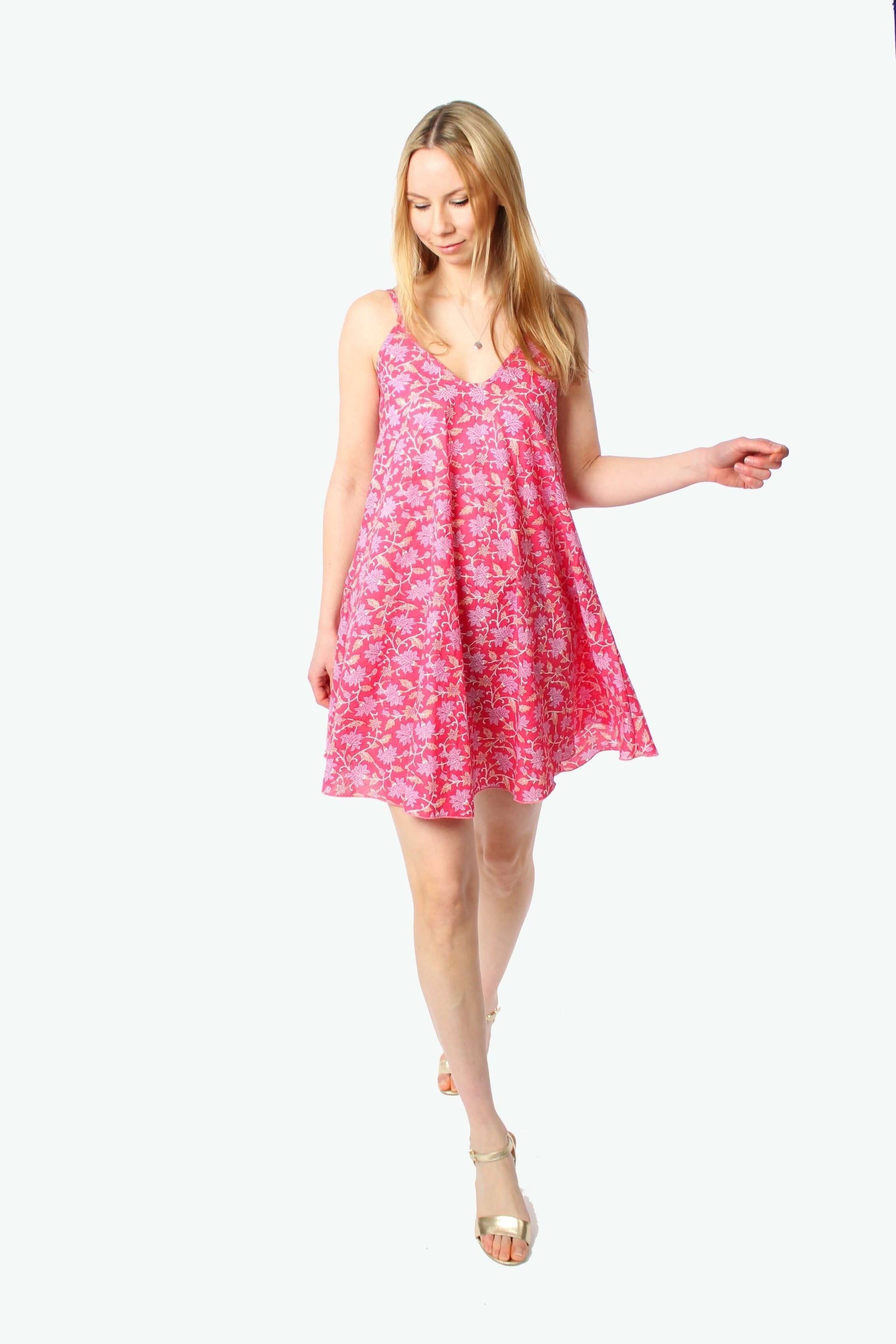 Ganzkörperaufnahme eines sommerlichen Trägerkleids in Pink mit floralem Muster, Minilänge, lockerer Schnitt, kombiniert mit goldenen Sandalen, freigestellt vor weißem Hintergrund.