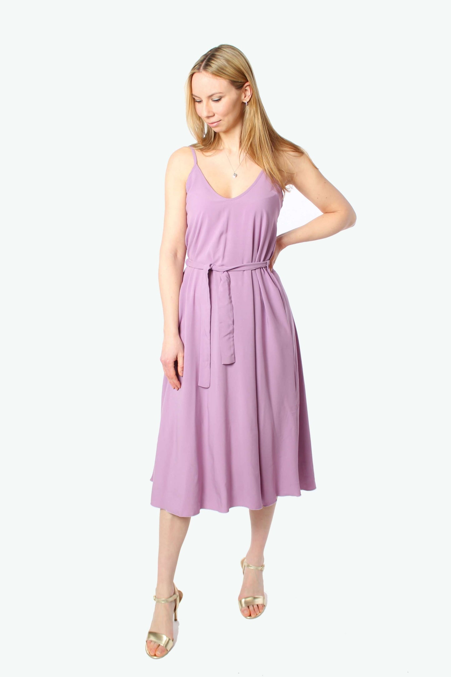 Blondes Model trägt ein fliederfarbenes Midikleid mit Spaghettiträgern und V-Ausschnitt, in der Taille mit einem fliederfarbenen Stoffgürtel gebunden, kombiniert mit goldenen Sandalen.