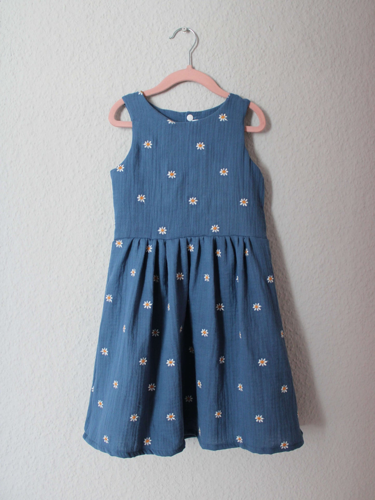 Kinderkleid in jeansblau mit weißen Gänseblümchen aus Musselinstoff von LA.LUKA Berlin auf einem Kleiderbügel in der Vorderansicht.